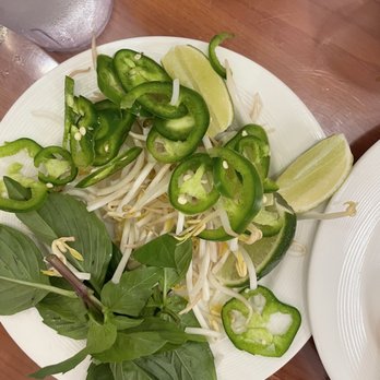 Pho 102 - Greens - The image shows a white plate filled with fresh ingredients including green herbs, bean sprouts, lime wedges, and sliced green chili peppers. The herbs appear to be basil, and the lime wedges are cut into quarters. The ingredients are arranged on a wooden table, likely served as accompaniments for a Vietnamese dish such as pho. - Vietnamese Near Me - Chantilly, Virginia