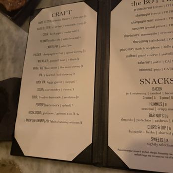 Spirited - Menu. - The image shows an open menu featuring sections for craft beers, cocktails, and snacks. The menu is placed on a marble table surface, with a small dish containing food partially visible in the top left corner. The menu includes detailed listings of beverages such as hard seltzers, ciders, pilsners, and IPAs, as well as snack options like bacon, hummus, and bar nuts. - Cocktail Bars Near Me - South Bend, Indiana