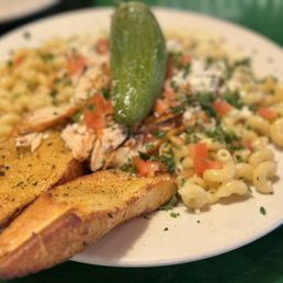 Alfredo the Dark - A white plate contains cooked macaroni pasta with chopped tomatoes, herbs, and pieces of grilled chicken on top. Two slices of garlic bread are placed at the edge of the plate, and a pickle is positioned upright in the center. The dish appears to be served in a casual dining setting, with a green table surface visible in the background.