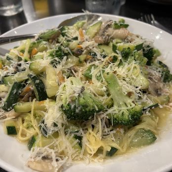 Clara's - Angel hair pasta with vegetable primavera sauce - A white bowl contains vegetable primavera pasta with cooked broccoli, zucchini, and shredded cheese on top. The dish appears freshly prepared and is served in a restaurant environment, with glasses and utensils visible in the background. - Italian Near Me - Woodridge, Illinois