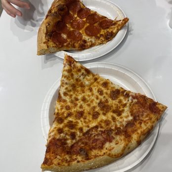 Costco - Two slices of pizza, one pepperoni and one cheese, are served on white disposable plates on a white table. A person wearing a gray shirt is partially visible, with their hand near the pepperoni slice. The setting appears to be casual, possibly in a pizzeria or a food court. - Wholesale Stores Near Me - Chicago, Illinois