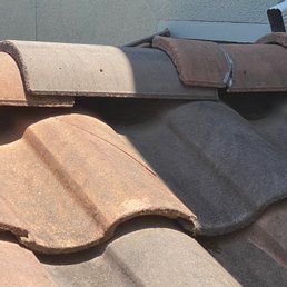 Tried to cover with the glue - The image depicts a close-up view of roof tiles made of clay or concrete, showing various colors including reddish-brown, gray, and dark brown. Some tiles appear weathered, with visible cracks and uneven surfaces. A section of the roof has been repaired or glued, as indicated by a visible adhesive or sealant applied to the tiles. The background includes part of a wall and a white gutter. The lighting suggests sunlight casting shadows on the tiles.