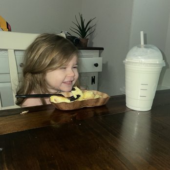 Starbucks - Babygirl w her bacon egg bites and her milk w whip! - A young girl with light brown hair is seated at a dark wooden table, smiling and looking at a dessert in a wooden dish, which appears to be a baked item with a spoon. Next to her is a large milkshake in a clear plastic cup with a straw and whipped cream on top. The background includes a white chair, a small table with a potted plant, and a wall with a picture frame. - Coffee & Tea Near Me - Willis, Texas