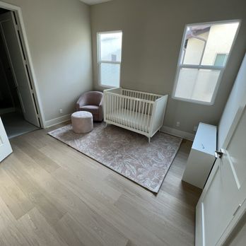 Divinity Flooring Services - Wood flooring - A nursery features a white crib, a pink armchair with a matching ottoman, and a patterned rug, with two windows providing natural light. - Flooring Near Me