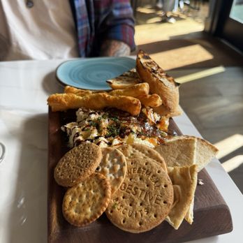 Thistledown & Row - Butter board - A cheese and cracker platter features various crackers, bread slices, and fries arranged on a wooden serving board. The platter includes a small blue plate nearby, with a person partially visible in the background, seated at a table in a restaurant setting with natural light coming through a window. - Cideries Near Me - Buchanan, Michigan