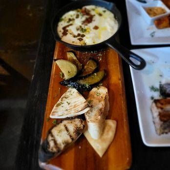 Debajo Tapas ABQ - Queso Flemeado - A rectangular wooden serving board displays grilled bread, roasted eggplant and zucchini slices, and a small black skillet containing a creamy white dip topped with herbs and spices. The presentation includes fresh herbs and lemon wedges, with additional dishes visible on white plates in the background, indicating a meal setting at a restaurant. - Spanish Near Me - Albuquerque, New Mexico