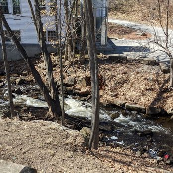 Station 101 - Sitting by the watee - A small stream flows through a wooded area with leafless trees and exposed soil. A building with white siding and windows is visible in the background, along with a sidewalk and steps leading up from the stream area. The scene appears to be in early spring or late fall, with no leaves on the trees and dry ground. - Beer Bar Near Me - Milford, New Hampshire