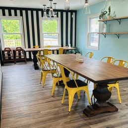 A bright dining room with a large wooden table surrounded by yellow metal chairs with black cushions. The room has a black-and-white striped accent wall with two windows, allowing natural light to enter. There are two wooden shelves with potted plants on the light blue wall, and modern pendant lights hanging from the ceiling. The flooring is wood laminate.