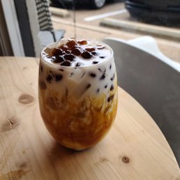 Brown sugar latte. - A glass of bubble tea with brown sugar syrup and tapioca pearls on top is placed on a light wooden table. The drink contains swirls of caramel-colored syrup and dark tapioca pearls. The background shows a window with parked cars outside, indicating the setting is likely a cafe or coffee shop. The table surface has some water stains or spills.