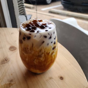 Soule' Coffee + Bubble tea - Brown sugar latte. - A glass of bubble tea with brown sugar syrup and tapioca pearls on top is placed on a light wooden table. The drink contains swirls of caramel-colored syrup and dark tapioca pearls. The background shows a window with parked cars outside, indicating the setting is likely a cafe or coffee shop. The table surface has some water stains or spills. - Coffee & Tea Near Me - Jackson, Mississippi