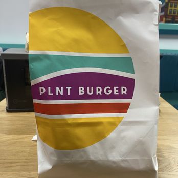 PLNT Burger - A paper bag featuring the PLNT Burger logo with a yellow, teal, purple, and red design, placed on a wooden table. - Fast Food Near Me - Brookline, Massachusetts