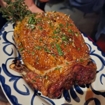 La Bodega - Duck (special 48hrs in advance order) - A large, cooked meat portion, possibly a steak or roast, is presented on a decorative plate with a blue and white pattern. The meat is garnished with chopped green herbs and appears glazed or sauced. The setting suggests a dining environment, with a hand and part of a person visible nearby. - Spanish Near Me - Watertown, Massachusetts