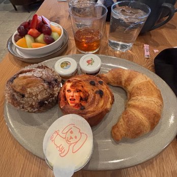 LUCIE drink + dine - Continental Breakfast - A breakfast setting on a wooden table includes a large oval plate with a croissant, a muffin decorated with a woman's face, and a pastry. Accompanying the plate are small containers of butter and jam. In the background, there is a bowl of mixed fruit, two glasses of water, a glass of iced tea, and several black mugs. The scene suggests a casual dining environment with a variety of breakfast items. - Bars Near Me - Boston, Massachusetts