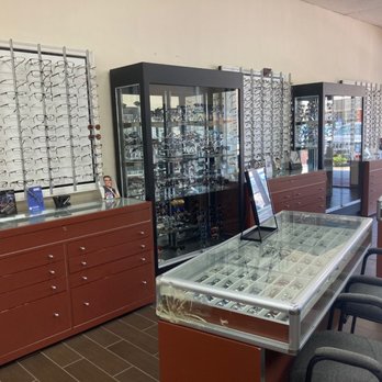 Eyeglass Express - Optical store interior featuring multiple wall-mounted eyeglass displays, glass cabinets filled with sunglasses, and seating for customers. The store has a window allowing natural light, and the displays include various styles of eyewear. The furniture includes wooden cabinets and chairs for customers. - Eyewear & Opticians Near Me - Temecula, California
