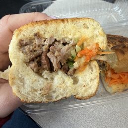 A close-up of a sandwich with sliced cooked beef, shredded carrots, and chopped celery inside a soft bread roll. The sandwich is held by a person's hand, with a plastic container in the background. The setting appears to be a casual dining or takeout environment.
