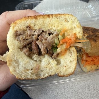 Pho Table - Bath Beach - A close-up of a sandwich with sliced cooked beef, shredded carrots, and chopped celery inside a soft bread roll. The sandwich is held by a person's hand, with a plastic container in the background. The setting appears to be a casual dining or takeout environment. - Vietnamese Near Me - Brooklyn, New York