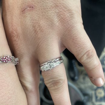 Benjamin - Recently cleaned wedding and engagement bands - The image depicts a close-up of a person's hand with three rings, including a wedding and engagement band, on the middle finger. The hand has a small healing scar or scratch near the knuckle. The background is blurred, showing part of a vehicle interior and a patterned floor mat. The rings are silver with one featuring multiple stones. The caption indicates the bands were recently cleaned. - Jewelry Near Me - Montrose, California