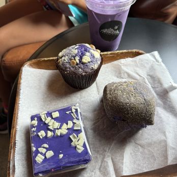Coffee Sarap - Ube brownie - A wooden tray lined with parchment paper displays three Filipino-inspired baked goods: a purple muffin topped with white chocolate chips, a purple cake with white chocolate shavings, and a purple bread roll. Behind the tray, a purple ube drink in a clear plastic cup with a straw is visible. The setting appears to be a cafe or bakery, with a person reading a comic book in the background. - Coffee & Tea Near Me - Denver, Colorado