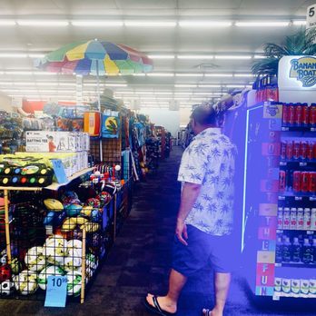 CVS Pharmacy - Sir Hubbers Flyin thru the CVS Universe yuge beachy section OuiOui.. - A man dressed in a white shirt with a palm tree pattern, dark shorts, and sandals is walking through an aisle in a retail store. The aisle contains beach-related products such as balls, umbrellas, and toys. Bright overhead lighting illuminates the store, and there is a colorful beach umbrella displayed above the products. The store appears to sell summer and recreational items, with shelves stocked with various beach accessories and toys. - Drugstores Near Me - St.Pete Beach, Florida