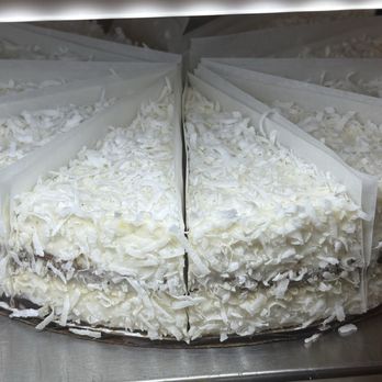 Bilqis Coffee - Coconut cake - A coconut cake with shredded coconut topping is shown, with a portion cut out, revealing the cake's interior. The cake is placed on a metal surface, likely in a bakery or cafe environment. The shredded coconut decorates the top and sides of the cake, which appears to be layered and frosted with white icing. - Coffee & Tea Near Me - Brooklyn, New York