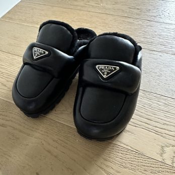 Prada - Mom's new furry plush shoes - A pair of black Prada slippers with a Prada logo on the top are positioned on a light-colored wooden floor. The slippers appear to be made of leather or a similar material, with a plush lining visible inside. The image captures the slippers from a slightly elevated angle, showing their design and branding clearly. - Accessories Near Me - McLean, Virginia