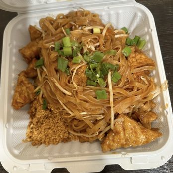 Royyim Thai - Vegan Vegan - Pad Thai - A disposable white takeout container contains Pad Thai noodles with a brown sauce, garnished with chopped green onions. There are pieces of chicken and a portion of crushed peanuts in the container. The container is open, revealing the food inside, and is placed on a dark wooden surface. - Thai Near Me - Mesa, Arizona