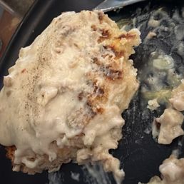 Biscuits and gravy, delicious! - A close-up image shows a serving of biscuits and gravy on a black plate. The biscuits are covered with a thick, creamy white gravy, with some parts of the biscuit visible underneath. The gravy appears smooth and slightly glossy. A spoon is partially visible, resting on the plate, with some gravy on it. The background includes a blurred view of a kitchen or dining area.