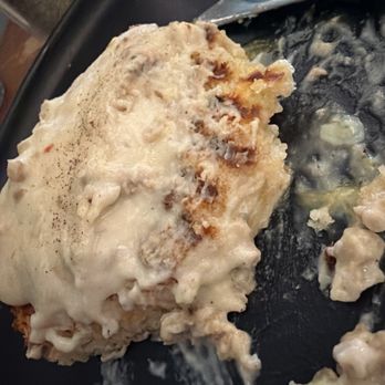 Jules' Bistro - Biscuits and gravy, delicious! - A close-up image shows a serving of biscuits and gravy on a black plate. The biscuits are covered with a thick, creamy white gravy, with some parts of the biscuit visible underneath. The gravy appears smooth and slightly glossy. A spoon is partially visible, resting on the plate, with some gravy on it. The background includes a blurred view of a kitchen or dining area. - New American Near Me - St. Cloud, Minnesota
