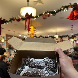 Traditional Ricotta - A person holds a box of cannoli pastries topped with powdered sugar and chocolate sprinkles inside a restaurant decorated for Christmas.