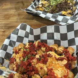 Spicy Chorizo Tater Tots - Baskets of loaded nachos and tacos with toppings like cheese, meat, guacamole, and salsa are placed on a wooden table in a casual dining setting.
