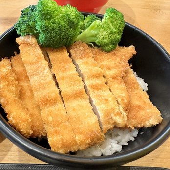 Bubblebee X Ramen - Chicken Katsu Rice Bowl - A black bowl filled with white rice is topped with several slices of breaded and fried cutlet, accompanied by a few pieces of steamed broccoli. The dish is placed on a wooden table, with a red bowl of similar food visible in the background. The presentation emphasizes the crispy texture of the cutlet and the vibrant green of the broccoli. - Ramen Near Me - Ashburn, Virginia