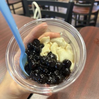 Scooptopia - Taho - A person holds a clear plastic cup filled with black tapioca pearls and white jelly pieces, with a blue straw inserted. The background shows a casual dining area with wooden tables and black chairs, indicating a food or dessert shop environment. - Ice Cream & Frozen Yogurt Near Me - Phoenix, Arizona