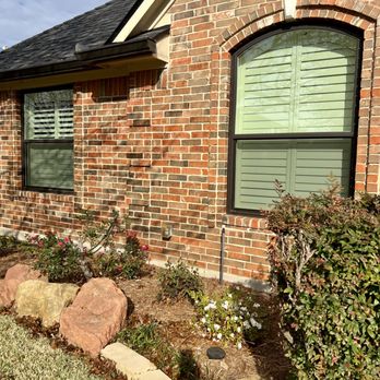 High Performance Roofing - A brick residential house with two large windows, each with green shutters, is visible. The house has a sloped roof with dark shingles and a small overhang. In front of the house, there is a landscaped garden with rocks, bushes, and flowering plants, bordered by a curved concrete edge. The scene is illuminated by sunlight, with a partly cloudy sky in the background. - Roofing Near Me - Frisco, Texas
