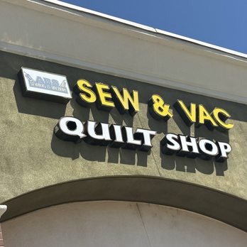 In Stitches - A storefront sign on a building exterior displays the business name 'SEW & VAC' and 'QUILT SHOP' in bold yellow and white letters. The sign is mounted on a textured green wall with a small logo or sign above it. The sky is clear and blue, and part of the building's roofline is visible. - Fabric Stores Near Me - Dixon, California