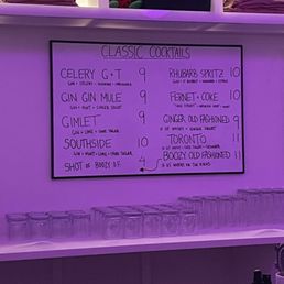 A handwritten menu board displays classic cocktails with prices, including gin, whiskey, and non-alcoholic options, in a bar setting with purple lighting.
