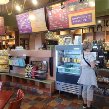 Ridley's Bakery Cafe - Pickup & Cooler Counter - A woman stands at the counter of Ridley's Bakery Cafe, looking at the menu screens above the service area, with a refrigerated display case nearby. - Bakeries Near Me - Troy, Michigan