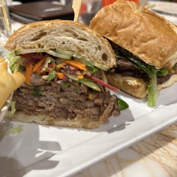 Biscotti's - Bahn mi - A close-up of a sliced beef sandwich with coleslaw, shredded carrots, and cucumbers on a toasted bun. The sandwich is placed on a white plate, with a second sandwich behind it. The background shows a restaurant setting with drinks and menus. The sandwich features thinly sliced beef, fresh vegetables, and a soft bun, typical of a deli or casual dining restaurant. - Cafes Near Me - Reno, Nevada