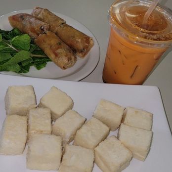Pho Leo - The image features a white rectangular plate with evenly cut cubed bread pieces. Behind it, there is a round white plate containing two spring rolls and a small bunch of mint leaves. To the right, a clear plastic cup filled with iced tea, ice cubes, and a straw is visible. The setting appears to be a casual dining environment, with a plain light-colored table surface. - Vietnamese Near Me - Mesa, Arizona