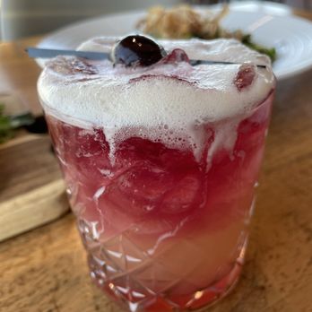 Devil's Kitchen - New York Sour - A pinkish-red cocktail with a foamy top and a cherry garnish is presented in a textured glass. The drink contains ice and appears to be served in a restaurant setting, with a blurred background of chairs and plates. The foam and cherry add visual appeal to the beverage. - New American Near Me - Grand Junction, Colorado
