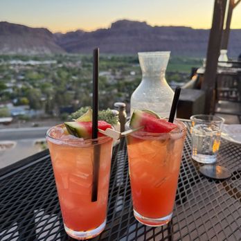 Sunset Grill - Drink special on the night of our visit was a watermelon margarita and they were great. - Two watermelon margaritas with watermelon and lime garnishes sit on a table overlooking a mountainous landscape at sunset. - Steakhouses Near Me - Moab, Utah