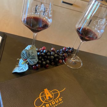 Aridus Tasting Room - Willcox - BS Merlot - The image shows two wine glasses filled with red wine on a dark table, with a decorative cluster of grapes and a leaf between them. A menu from Aridus Wine Company is also on the table. The background features a wooden counter with wine bottles and a light-colored wall, indicating an indoor wine tasting room setting. - Wineries Near Me - Willcox, Arizona
