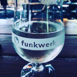 A transparent glass with the 'funkwerks' logo contains a clear beverage, set on a wooden table with a blurred outdoor background.