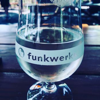 Funkwerks - A transparent glass with the 'funkwerks' logo contains a clear beverage, set on a wooden table with a blurred outdoor background. - Breweries Near Me - Fort Collins, Colorado