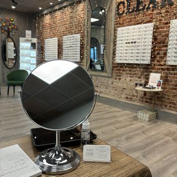ClearView Optical - My visit - Inside an optical shop, display boards with eyeglasses are mounted on a brick wall, with mirrors and a seating area visible. A table with a mirror, business card, and other items is in the foreground. - Eyewear & Opticians Near Me - Gainesville, Georgia
