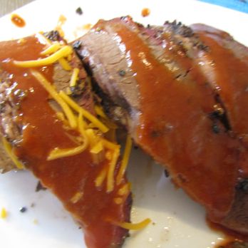 Lum's - Leftovers - Sliced Brisket atop Baked Potato - at Home - - Slices of smoked brisket with a dark, smoky exterior and pink smoke ring are arranged on a white plate. The meat is topped with barbecue sauce and shredded cheddar cheese. The background includes a blue surface, indicating a casual dining environment, likely at a barbecue restaurant. - Barbeque Near Me - Junction, Texas