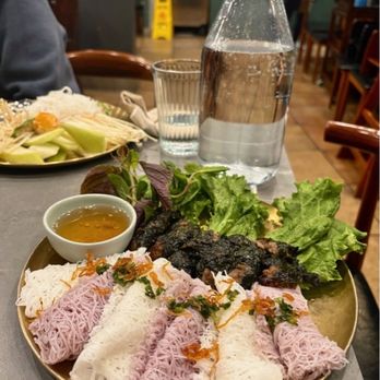 Bacogai - A plate of Vietnamese cuisine includes rice noodle rolls topped with herbs and garnishes, accompanied by grilled meat, fresh lettuce, and a small bowl of dipping sauce. The dish is presented on a round, metallic plate on a restaurant table, with a glass of water, a water pitcher, and another plate of food visible in the background. The setting appears to be a casual dining restaurant with wooden chairs and tables. - Hot Pot Near Me - Alameda, California
