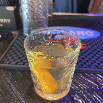 Devil's Hideaway - Old fashioned - A transparent glass with a decorative pattern holds a cocktail with a lemon wedge and ice cubes. The drink is on a dark textured bar counter, with a bar mat and a blurred background showing a person and bar equipment. - Wine Bars Near Me - Tempe, Arizona