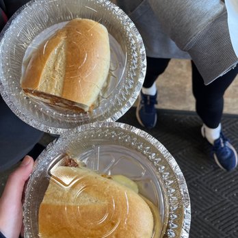 Nyack Diner - Two sandwiches in aluminum trays are held by two people, one in dark clothing and the other in gray, with a background of a person in gray sneakers and black leggings. - Breakfast & Brunch Near Me - Nyack, New York