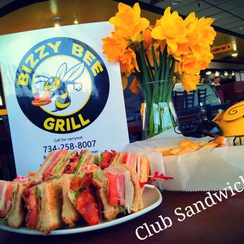 Bizzy Bee Grill - The image shows a table at Bizzy Bee Grill with a plate of club sandwiches featuring layers of bread, meats, and vegetables. A bottle of Frank's RedHot hot sauce is positioned to the left of the plate. Behind the food, there is a sign with the Bizzy Bee Grill logo, a cartoon bee, and contact information. A clear glass vase filled with yellow daffodils and bee-themed decorations, including a yellow bee figurine, are also on the table. The background reveals a restaurant interior with tables, chairs, and a few patrons. - American Near Me - Southgate, Michigan