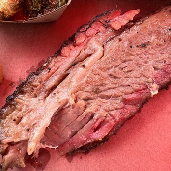 Truth BBQ - Brisket - A sliced smoked brisket with a dark, charred crust and pink smoke ring, placed on a red surface, with side dishes partially visible. - Barbeque Near Me - Brenham, Texas